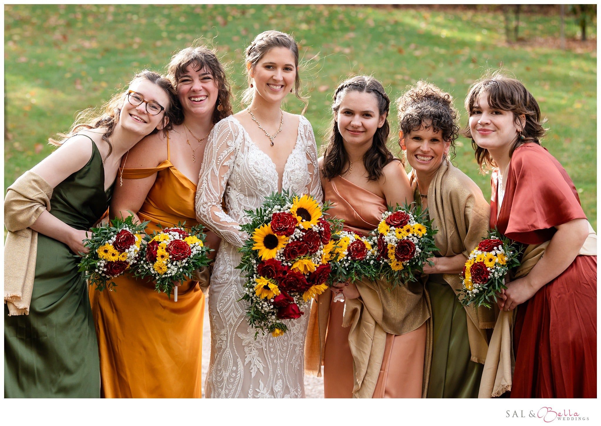 Bridesmaids in different fall-toned dresses posing with the bride while holding rose and sunflower bouquets. 