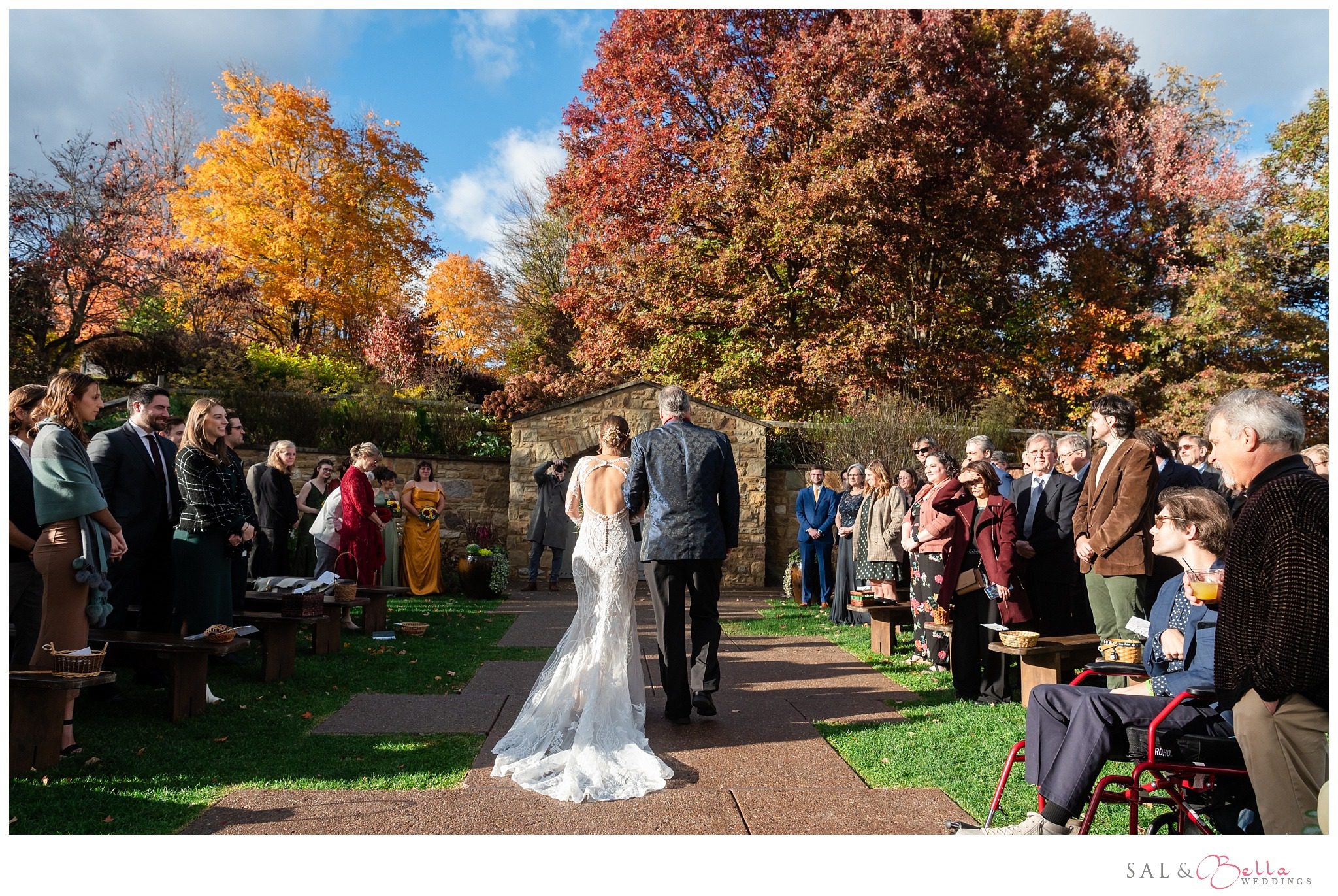 Bride walking down the aisle with her father, surrounded by autumn trees and floral decor.