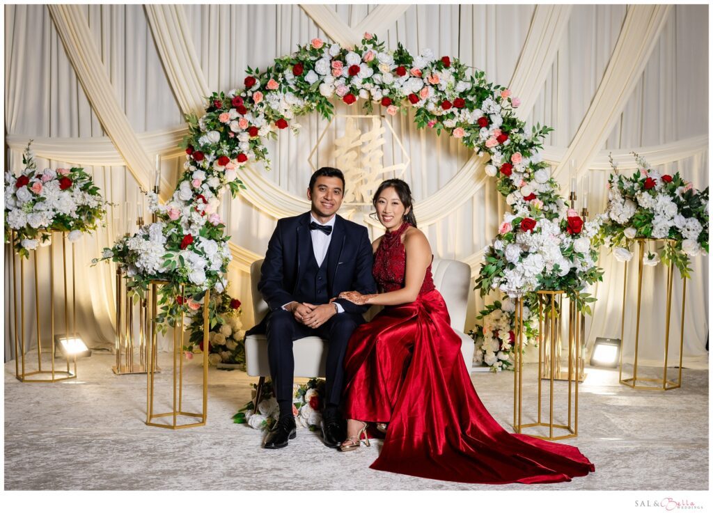 Beautiful draped mandap with floral arch featuring red pink and white flowers. Bride & groom pose on their chaise. Bride is wearing a red gown.