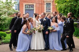 A wedding party is laughing in the courtyard garden at the Priory. Bridesmaids are wearing periwinkle-colored dresses.