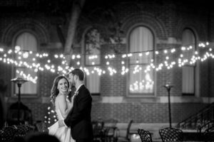 A couple poses for a photo in the courtyard of the priory at night with string lights in the background.