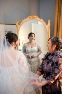 Brides mother fluffs her veil, you can see the brides expression in a full-length gold mirror in her Priory Hotel Suite.