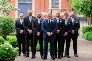 Groom poses for photos with his groomsmen the grand hall at the priory is in the background.